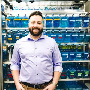 Alex Seaver stands in front of an array of little fish tanks wearing a purple button up shirt.