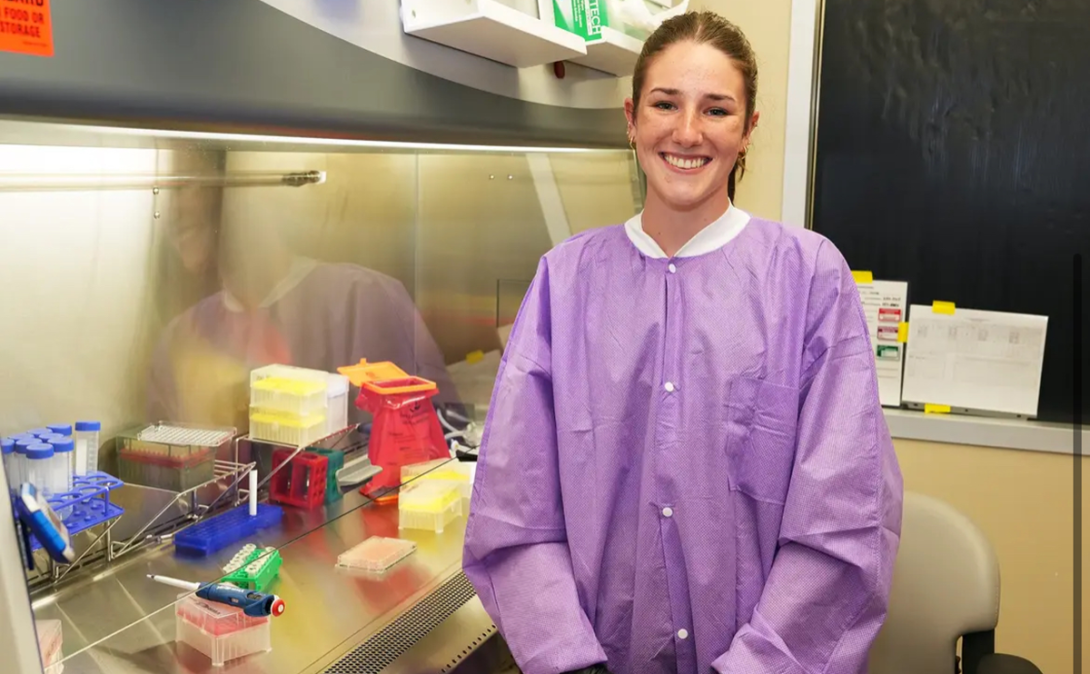Liliane Moorhead wearing a purple smock standing next to a lab hood with various instruments.